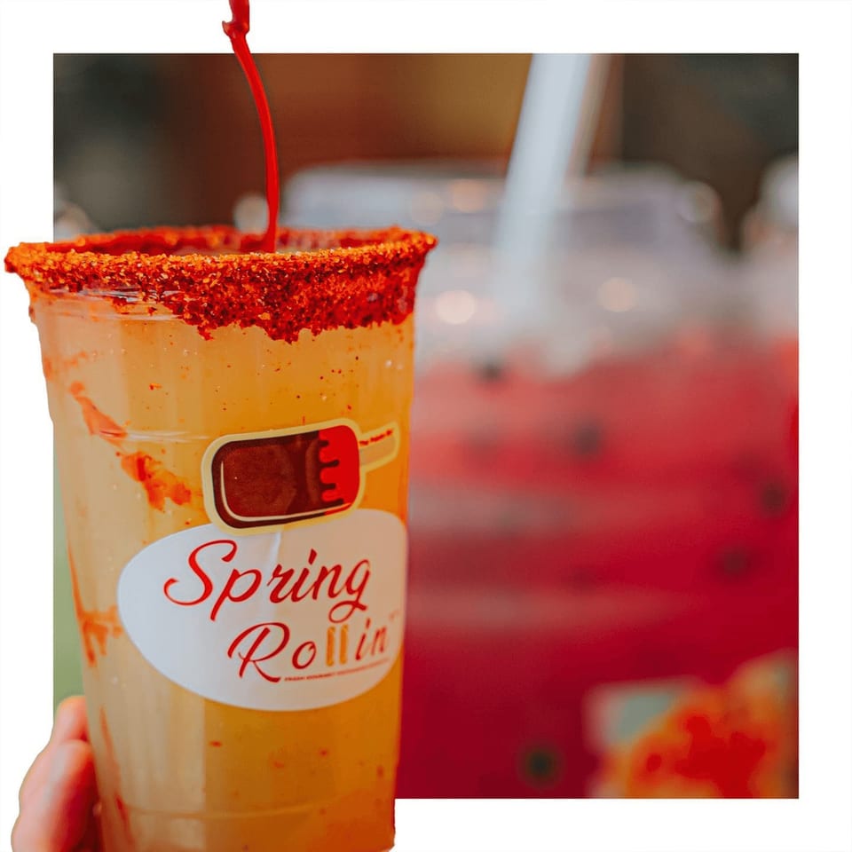 A hand holds a Spring Rollin' drink with a chili-rimmed cup and red liquid pouring in.