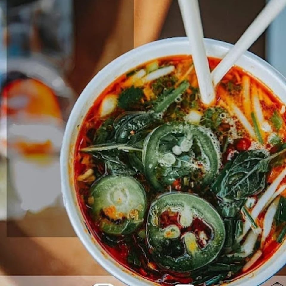 Close-up of a vibrant Vietnamese soup with green vegetables, red broth, and chopsticks.