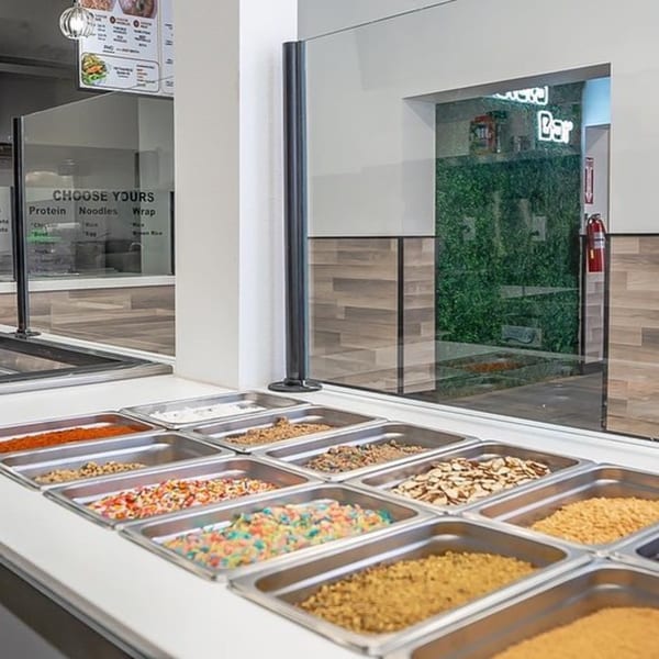 A Vietnamese restaurant's topping bar with various ingredients in metal trays, behind a sneeze guard.