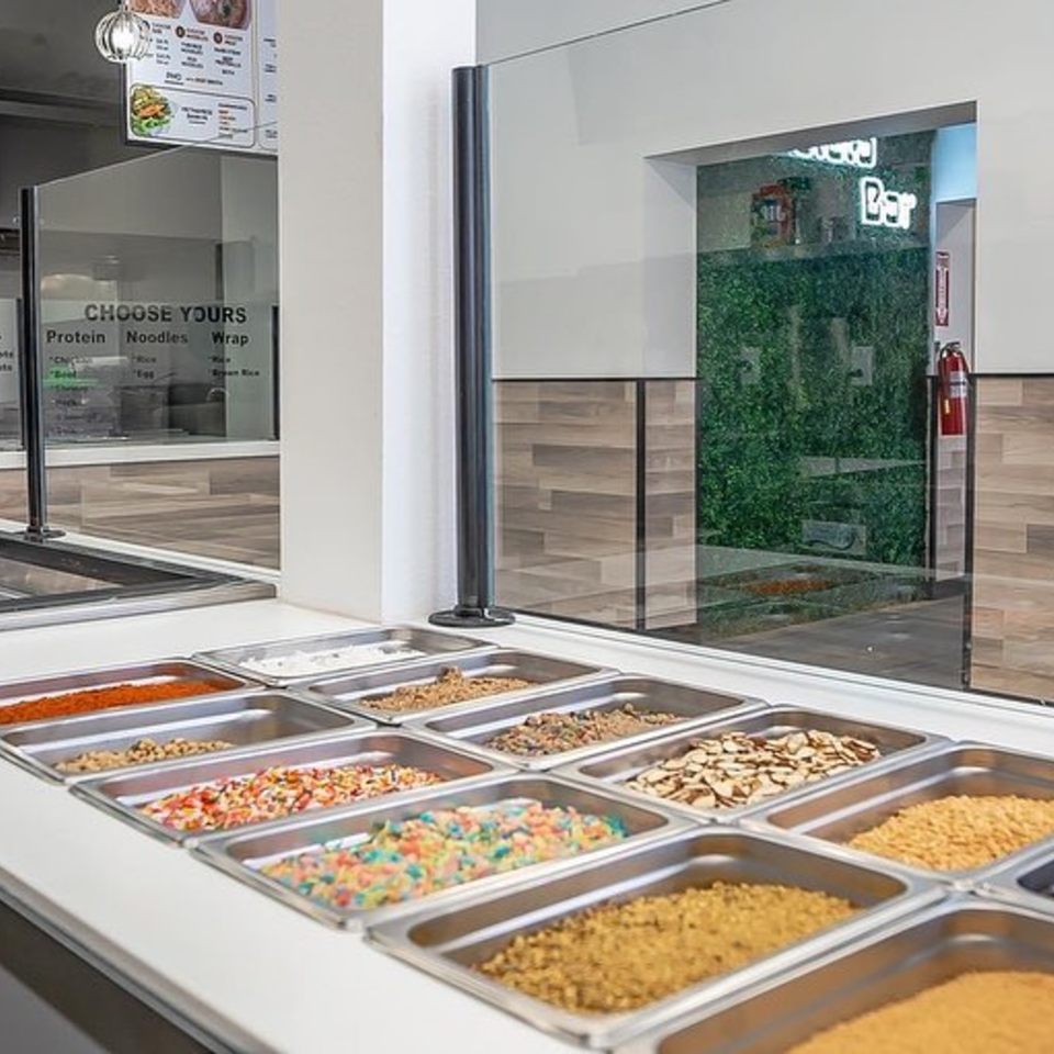 A Vietnamese restaurant's topping bar with various ingredients in metal trays, behind a sneeze guard.