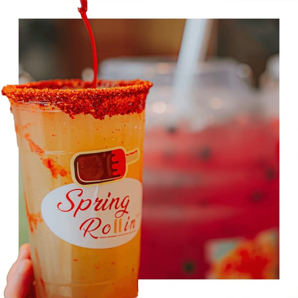 A hand holds a Spring Rollin drink with a chili-lime rim, red sauce pouring in.