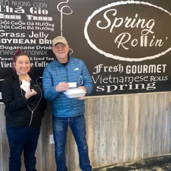 Two smiling people, a woman and a man, holding food containers and cards in front of a Spring Rollin' menu.
